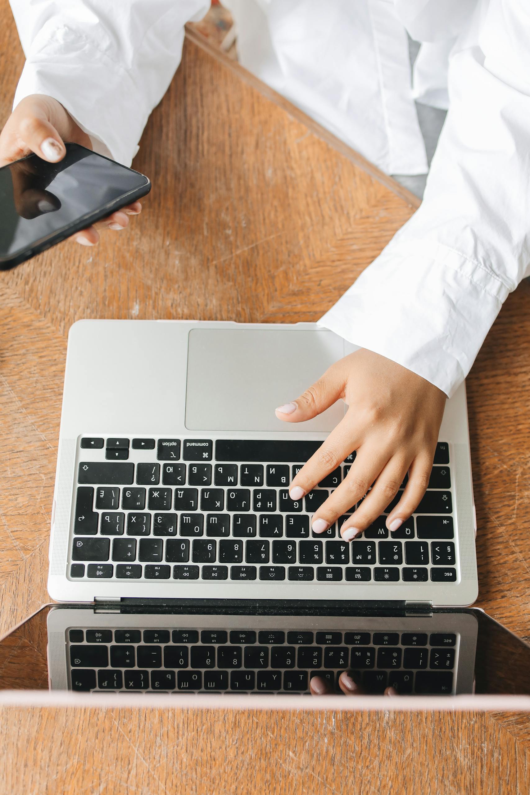 Overhead shot of hands using laptop and smartphone on wooden desk.